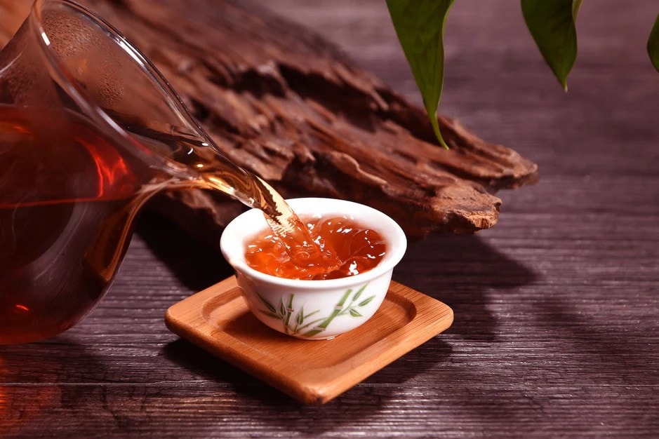 Dark amber tea is poured from a clear glass pitcher into a small, white ceramic cup decorated with bamboo motifs. The cup sits on a square wooden coaster against a rustic backdrop of weathered wood and green leaves.