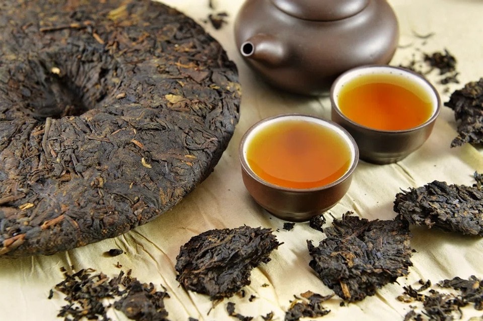 The image displays a large, compressed disk of dark Pu-erh tea alongside several broken-off chunks of loose tea leaves. Two small, brim-filled cups of amber liquid sit next to a matching clay teapot on a textured, light-colored surface.