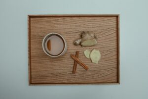 An overhead shot shows a wooden tray holding a white cup of tea, a piece of fresh ginger root with two slices, and two cinnamon sticks.