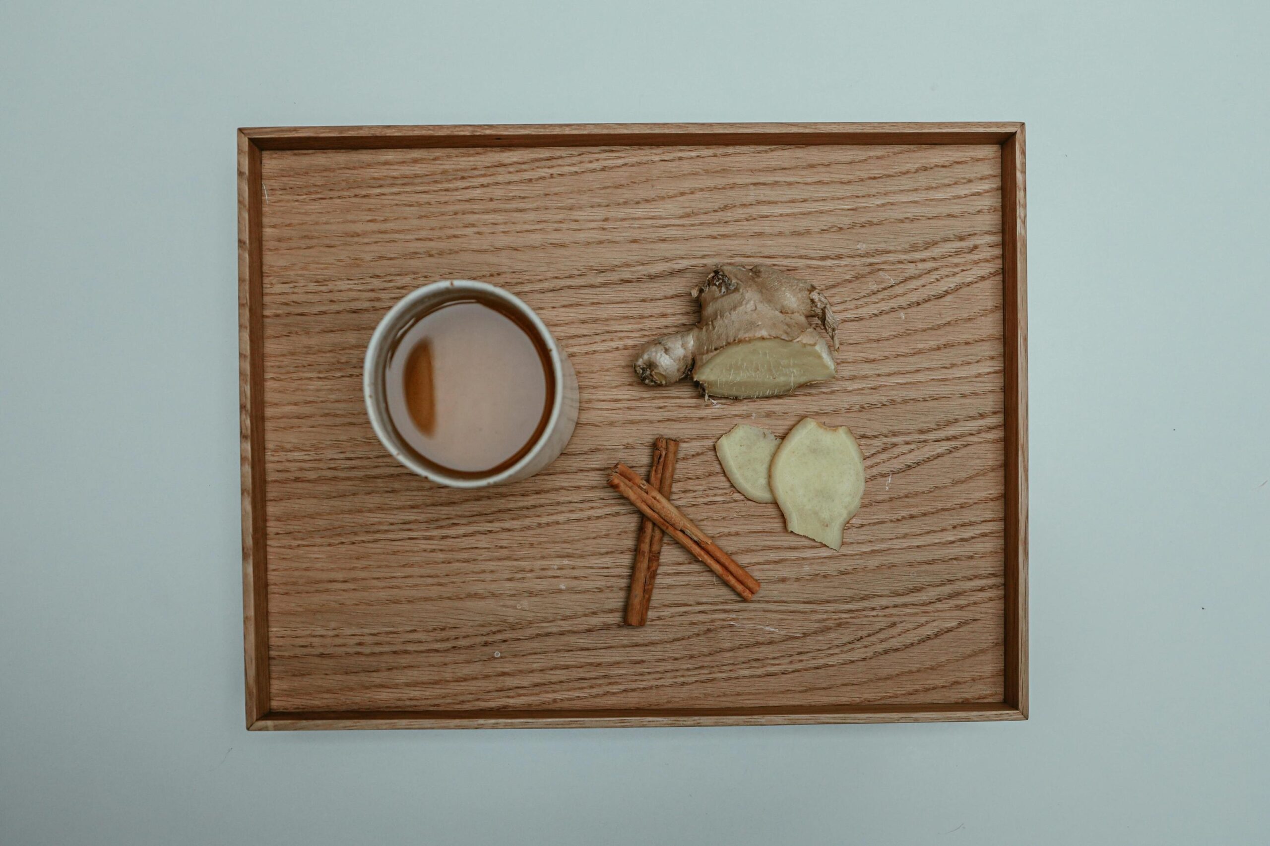 An overhead shot shows a wooden tray holding a white cup of tea, a piece of fresh ginger root with two slices, and two cinnamon sticks.