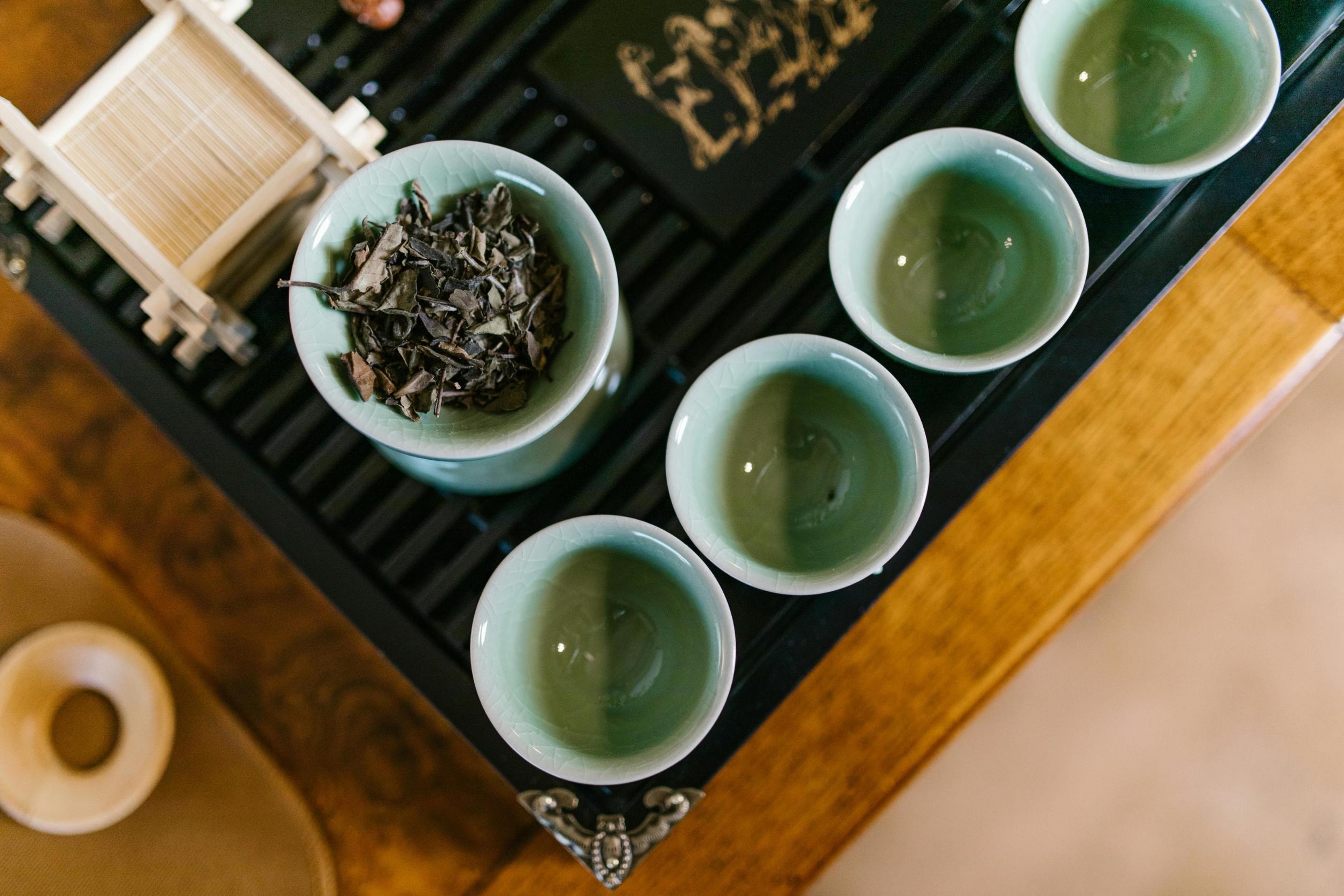 This image shows a top-down view of a traditional tea set featuring several small, light-green ceramic cups and a bowl of dried tea leaves. The items are neatly arranged on a dark, slatted tea tray set atop a warm wooden table.