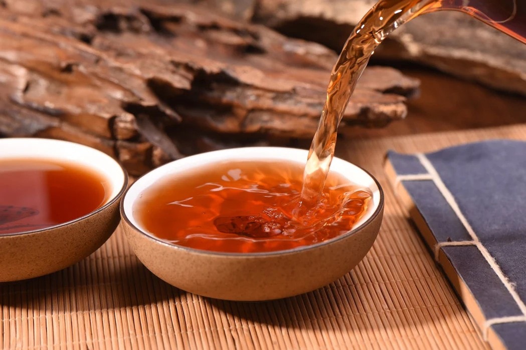 Amber-colored tea is poured from a glass vessel into a wide, ceramic cup resting on a traditional bamboo mat. In the background, a weathered piece of wood and a blue bound book add to the rustic, scholarly atmosphere of the scene.