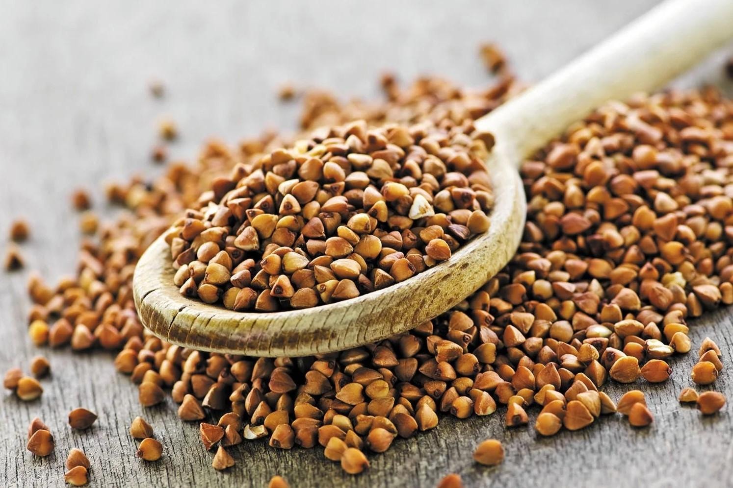 A wooden spoon overflows with toasted buckwheat groats, which are scattered across a rustic gray wooden surface. The triangular seeds are a rich, warm brown color and serve as the central focus of the close-up shot.