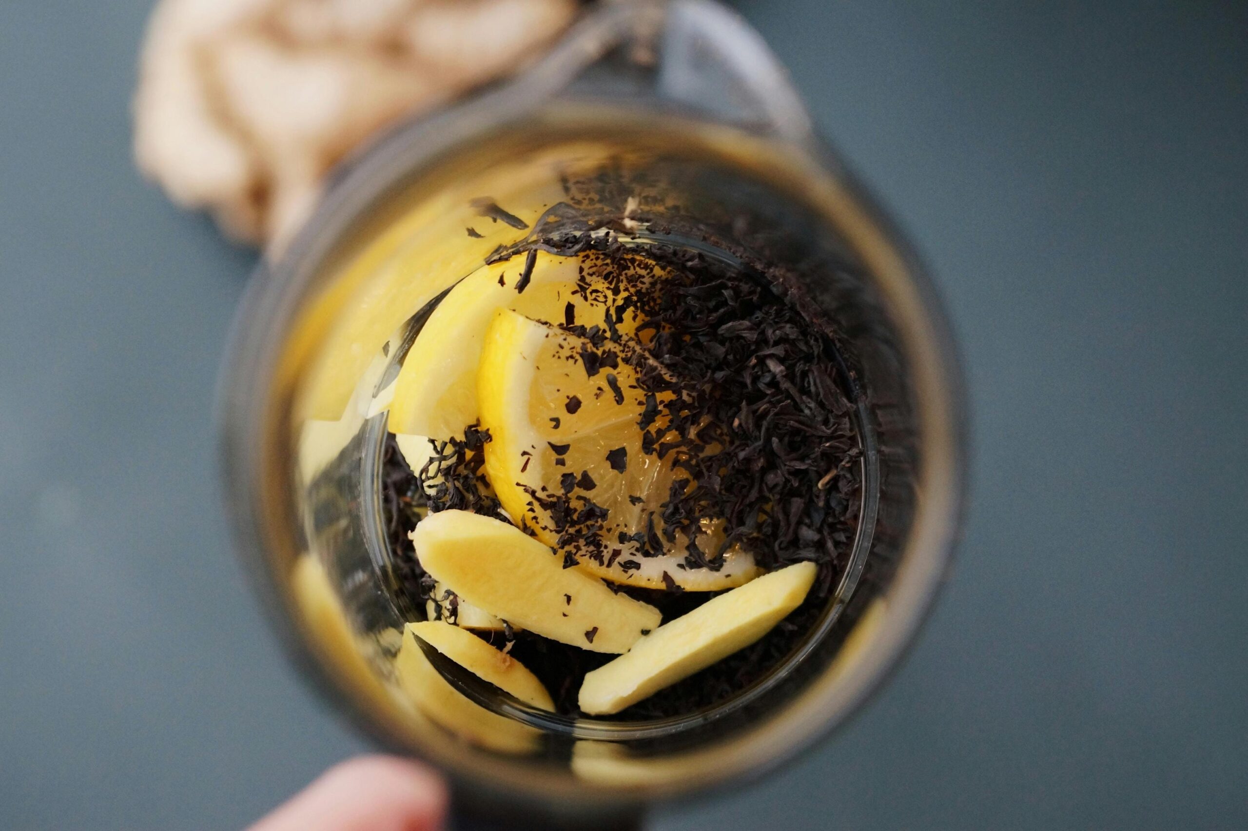 An overhead, close-up shot captures loose black tea leaves, fresh lemon slices, and pieces of ginger inside a glass teapot, ready to be brewed.