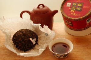 This image displays a dark, fermented Pu-erh tea Tuocha unwrapped on a wooden surface next to its red and gold decorative canister. Accompanying the tea is a traditional Yixing clay teapot and a small ceramic cup filled with deep amber-colored brewed tea.