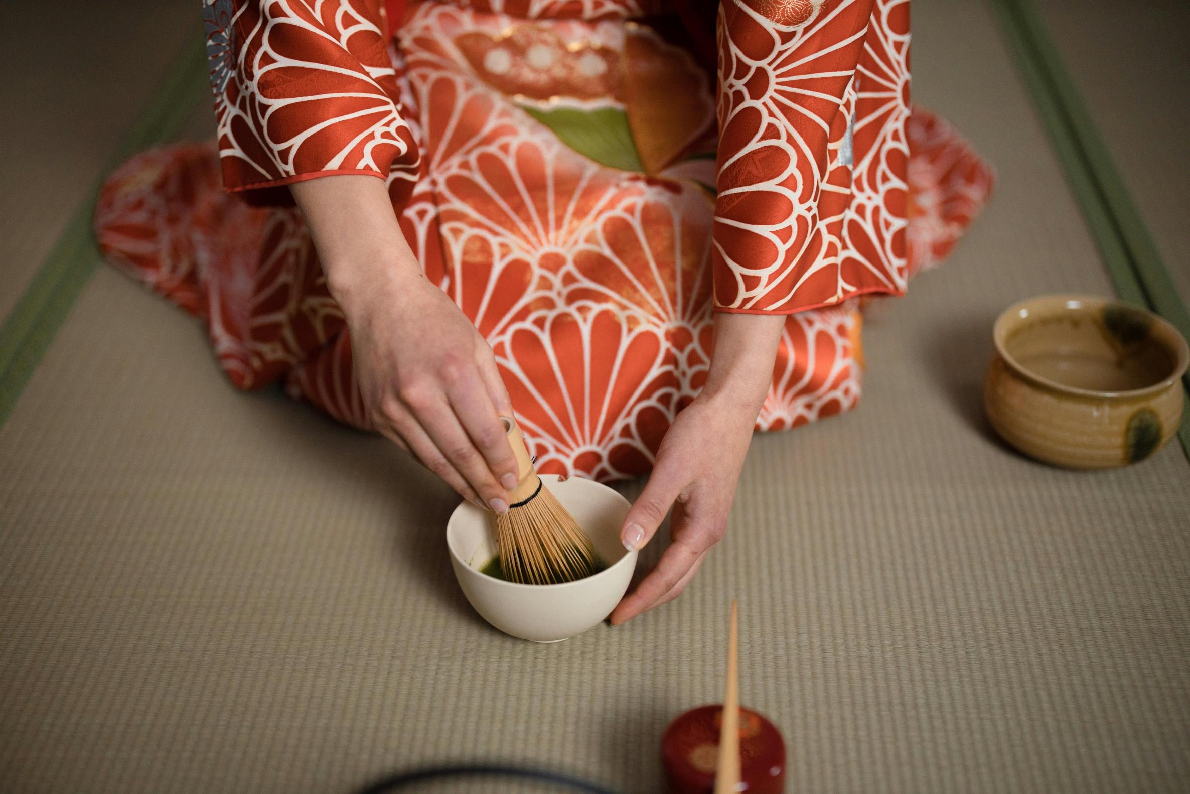 A person wearing a vibrant, floral-patterned kimono is seated on a tatami mat while whisking green matcha in a ceramic bowl. Surrounding them are traditional tea ceremony items, including a wooden scoop and a separate tea container.