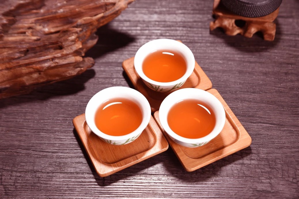 Three small, white ceramic cups filled with golden-amber tea are arranged on square wooden coasters. The scene is set on a dark, textured surface featuring a piece of rustic driftwood and green leaves in the background.
