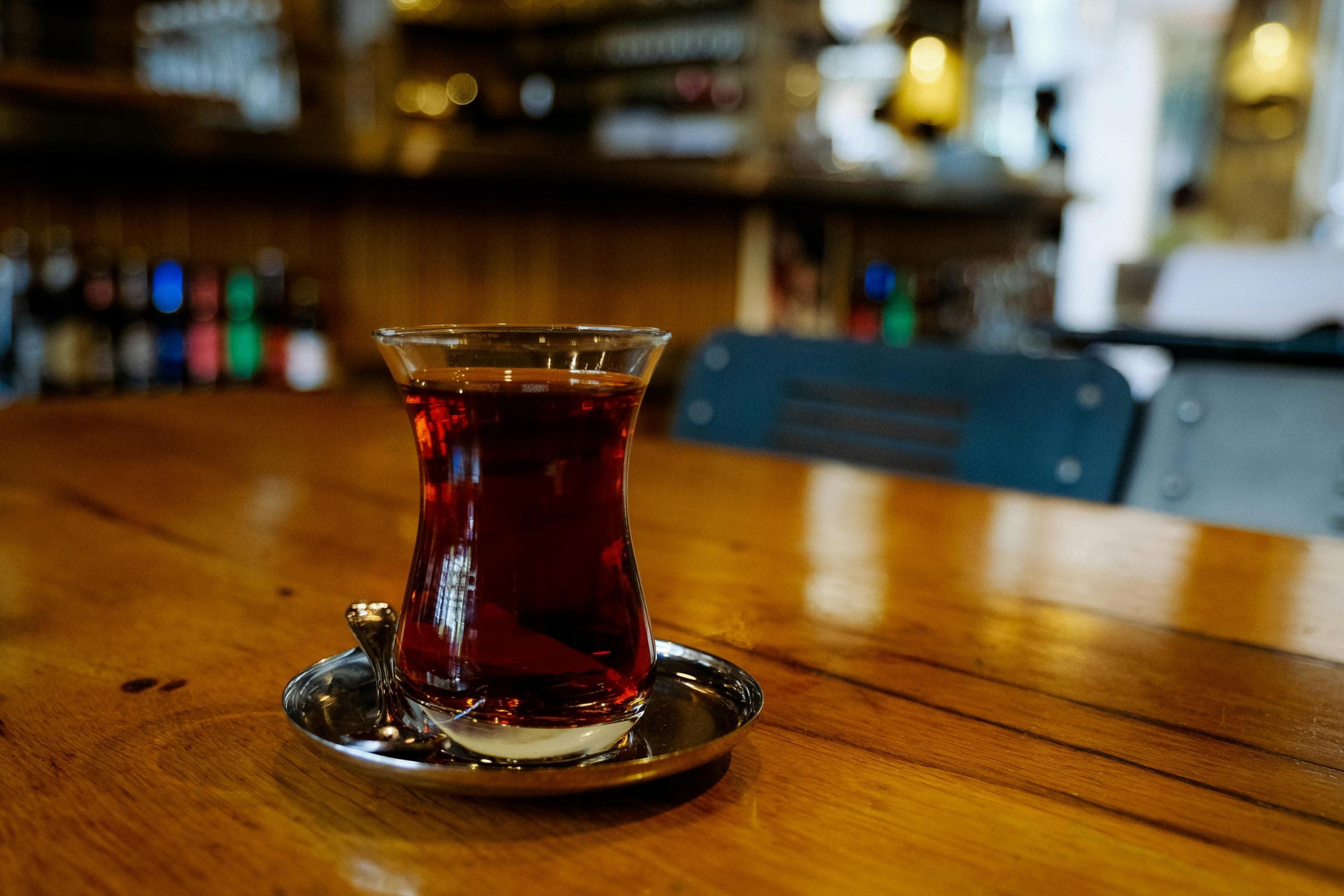  A tulip-shaped glass filled with dark, amber-colored tea sits on a small silver saucer atop a polished wooden table. In the background, the blurred interior of a cafe or bar creates a warm, cozy atmosphere with soft lighting and stacked bottles.