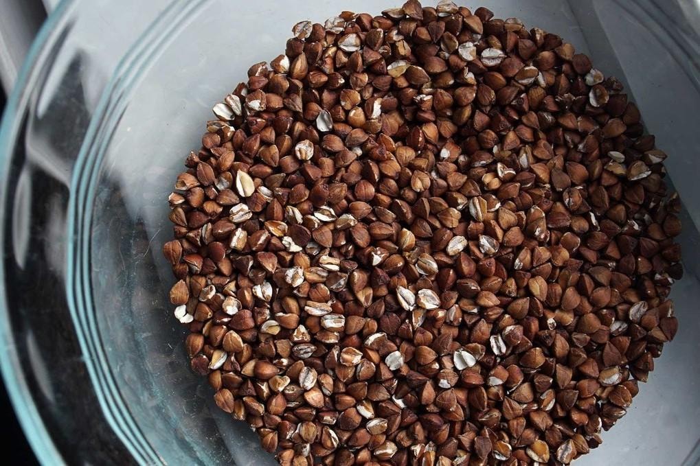 A top-down view shows a glass bowl filled with a large quantity of roasted, dark-brown buckwheat groats. The triangular grains are packed closely together, showing a mix of solid hulls and some pale, cracked interiors.