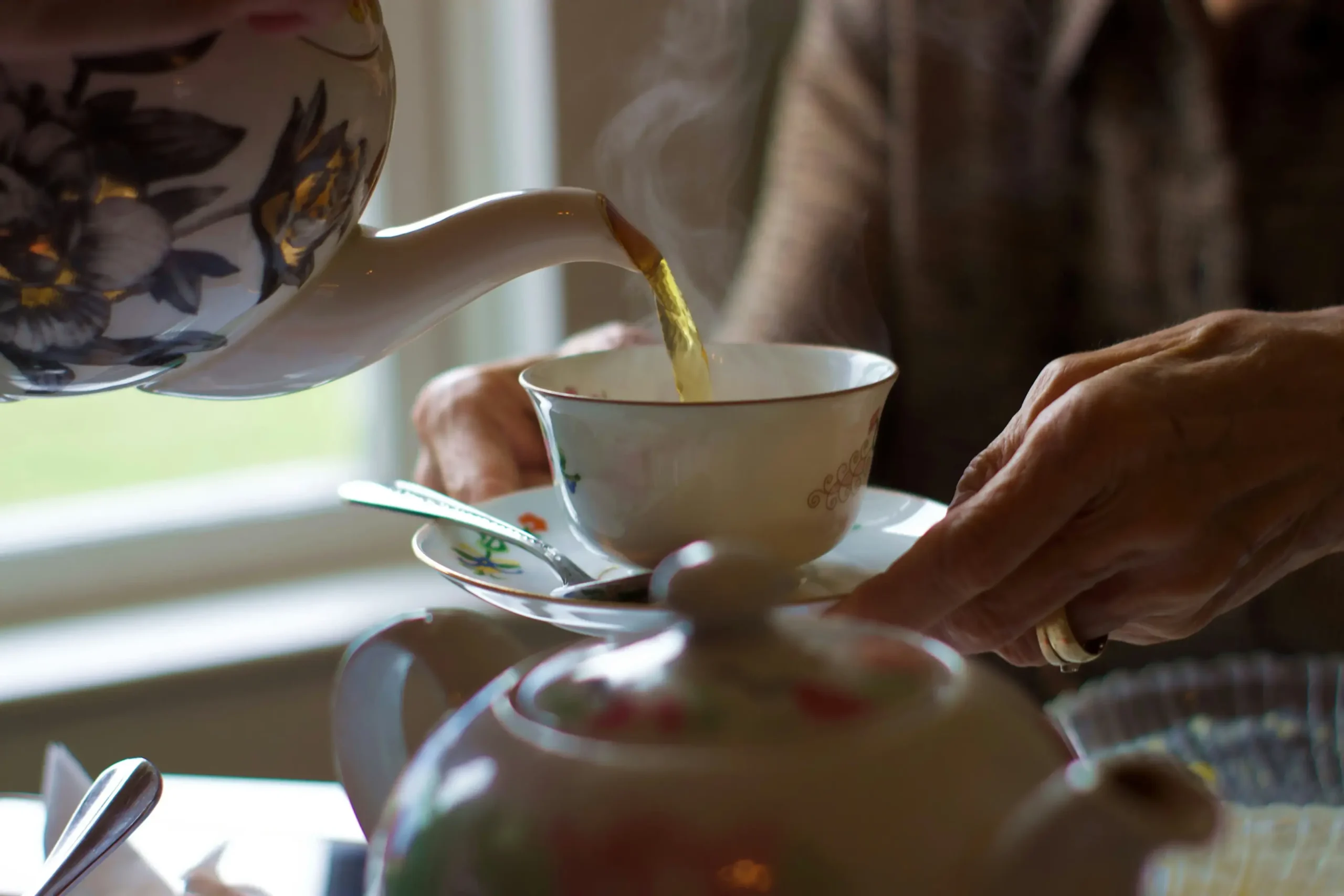 A person’s weathered hands carefully steady a delicate floral teacup and saucer while steaming tea is poured from a matching teapot. The scene captures a warm, intimate moment, highlighted by the soft light and rising vapor.