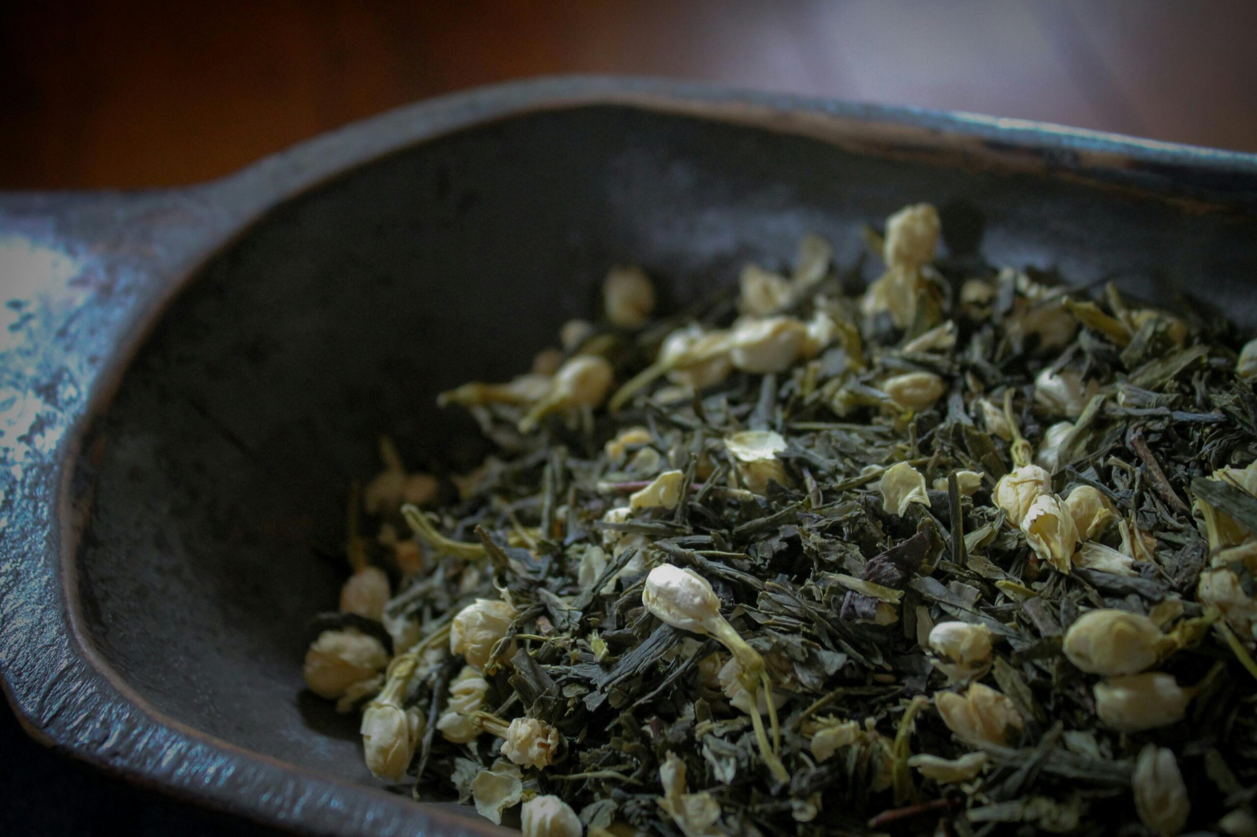 A rustic wooden bowl is filled with a mixture of dark green tea leaves and small, dried jasmine flower buds. The image features a shallow depth of field, highlighting the delicate textures of the botanicals against a dark, moody background.
