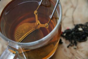 A mesh tea infuser is submerged in a clear glass mug filled with warm, amber-colored tea. Beside the mug, a small pile of loose-leaf tea rests on a textured, light-colored surface.