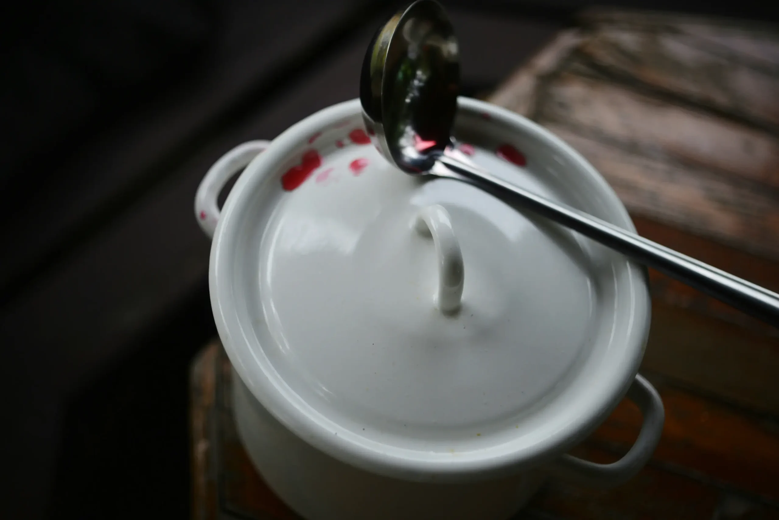 A white enamel pot with a matching lid sits on a rustic wooden surface, featuring small red stains around its rim. A stainless steel ladle rests across the top of the pot, catching a faint reflection in its polished surface.