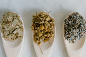 Three wooden spoons are arranged vertically, each holding a different heap of dried herbs and flower buds. The contents appear to be loose-leaf tea ingredients, including green leaves, yellow chamomile flowers, and purple lavender.