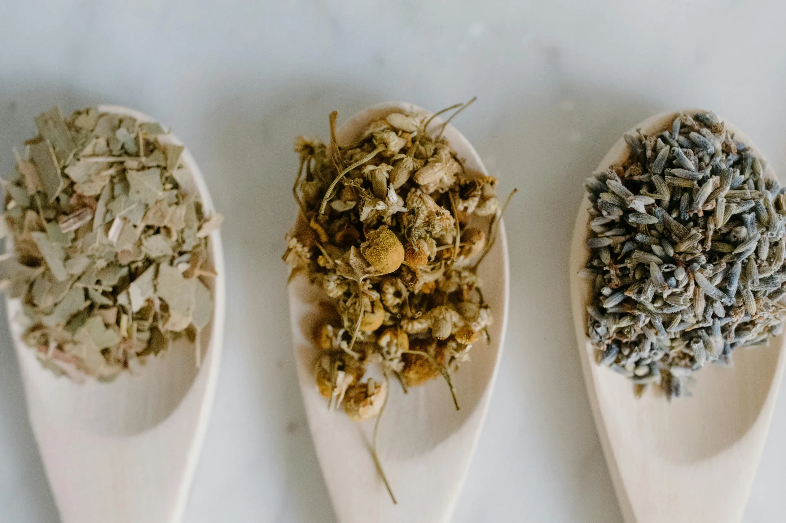 Three wooden spoons are arranged vertically, each holding a different heap of dried herbs and flower buds. The contents appear to be loose-leaf tea ingredients, including green leaves, yellow chamomile flowers, and purple lavender.