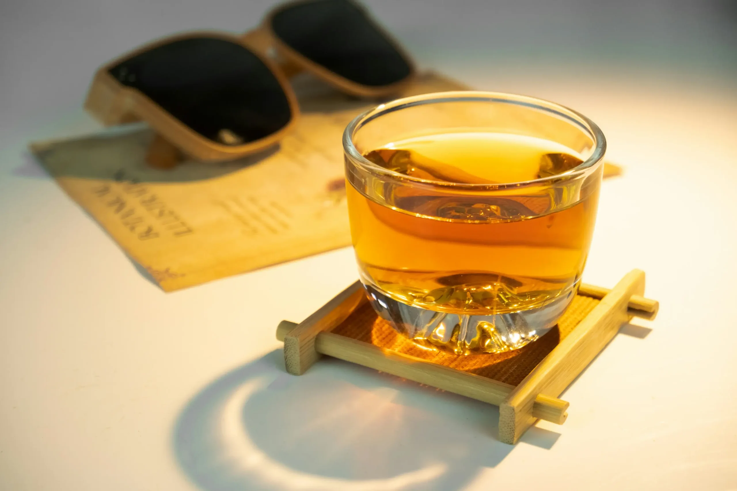 A small glass of amber-colored tea sits on a bamboo coaster, illuminated by a warm, golden light. In the soft-focus background, a pair of wooden sunglasses rests on a vintage botanical card.