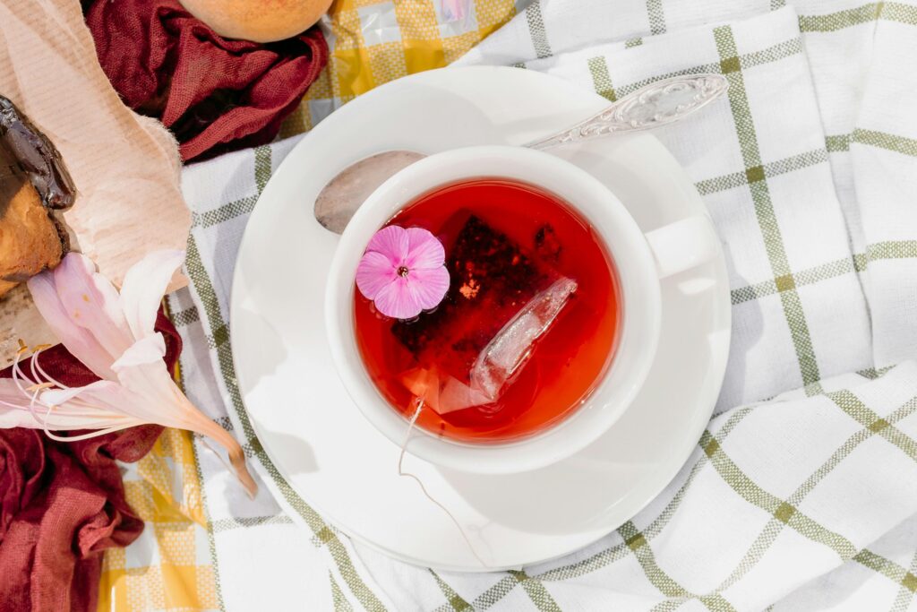 An overhead shot captures a white teacup filled with vibrant reddish-pink tea, garnished with a small purple flower and an submerged tea bag. The cup sits on a white and green checkered cloth, surrounded by a silver spoon, delicate flowers, and a glimpse of a chocolate pastry.
