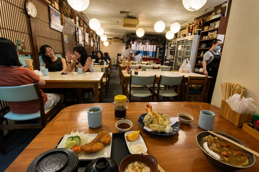 A warm and bustling Japanese restaurant interior is captured, featuring patrons seated at wooden tables under a series of spherical paper lanterns. The foreground showcases a table filled with a traditional meal, including a plate of fried fish with a side salad, a generous portion of shrimp tempura, a bowl of curry, and small cups of tea. In the background, other diners converse at their tables while a staff member wearing a face mask moves through the space, creating an inviting, authentic atmosphere for enjoying shared meals.