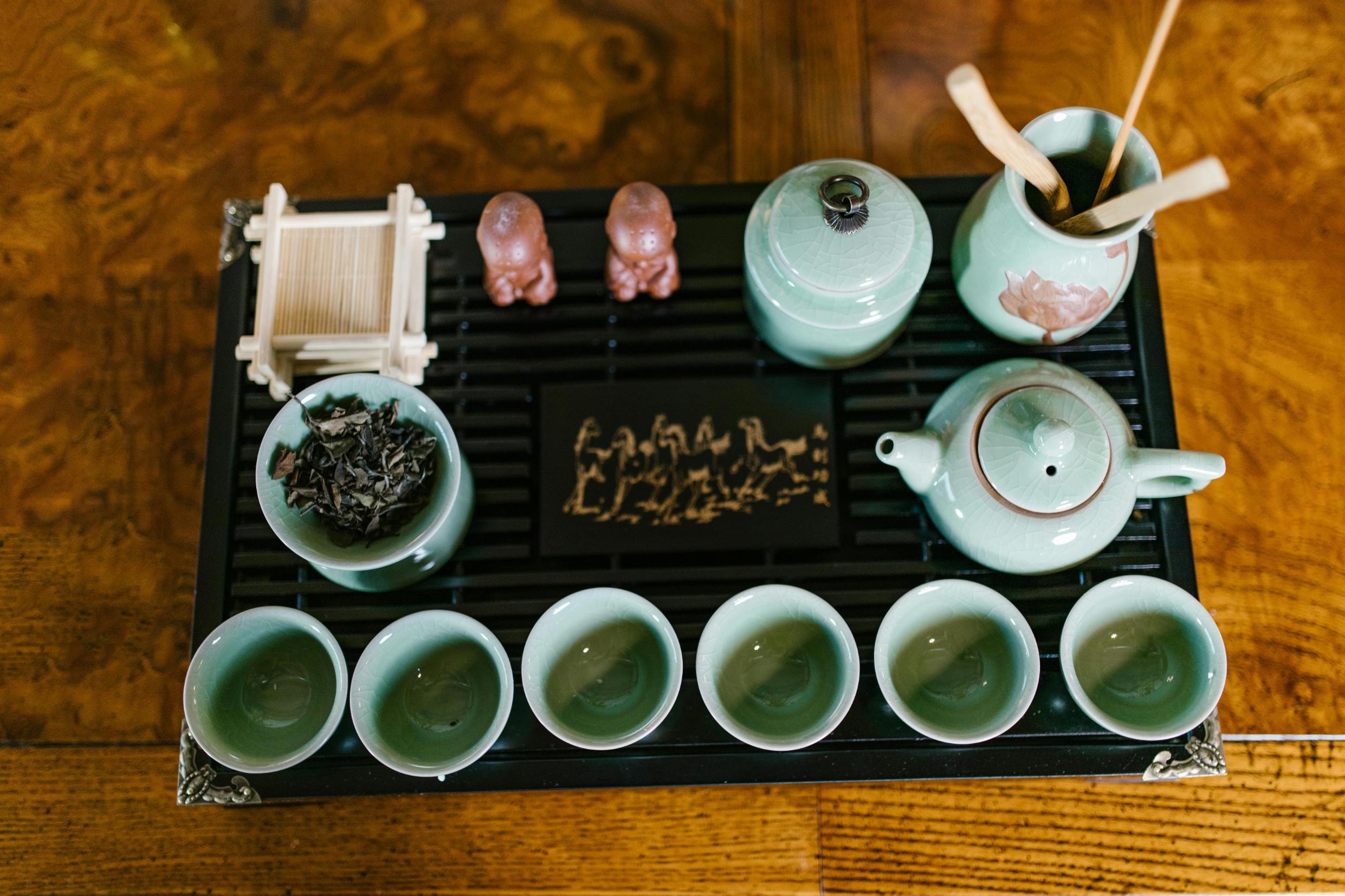 This top-down view features a light green celadon tea set, including six matching cups and a teapot, arranged on a black slatted tray. The set is accompanied by two small monk figurines, a tea canister, and wooden utensils, all set against a rich wood grain background.