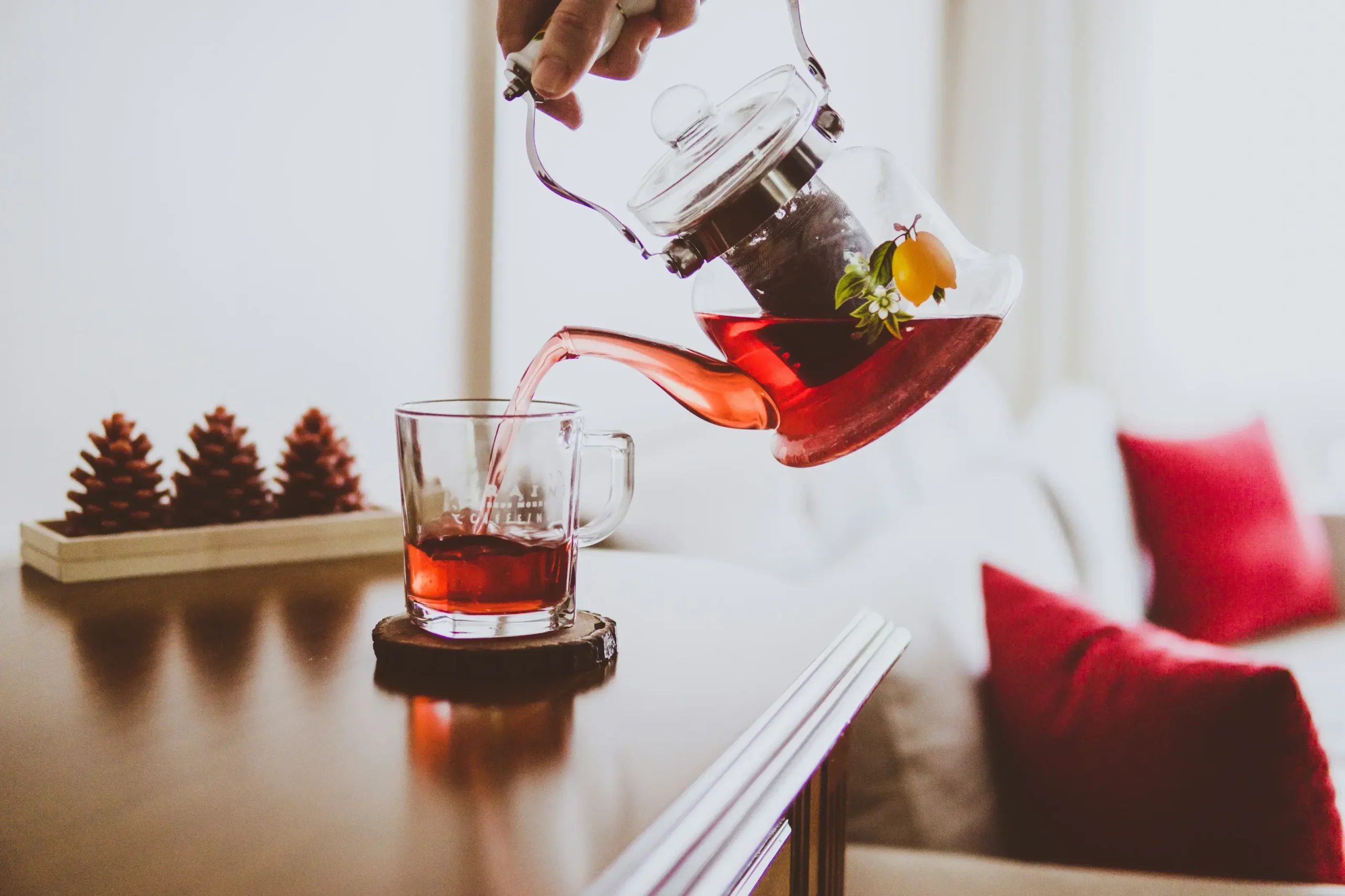 A hand pours a vibrant reddish tea from a clear glass teapot into a matching mug resting on a wooden table. The cozy indoor scene is accented by warm sunlight, red pillows on a couch, and small decorative pinecones in the background.