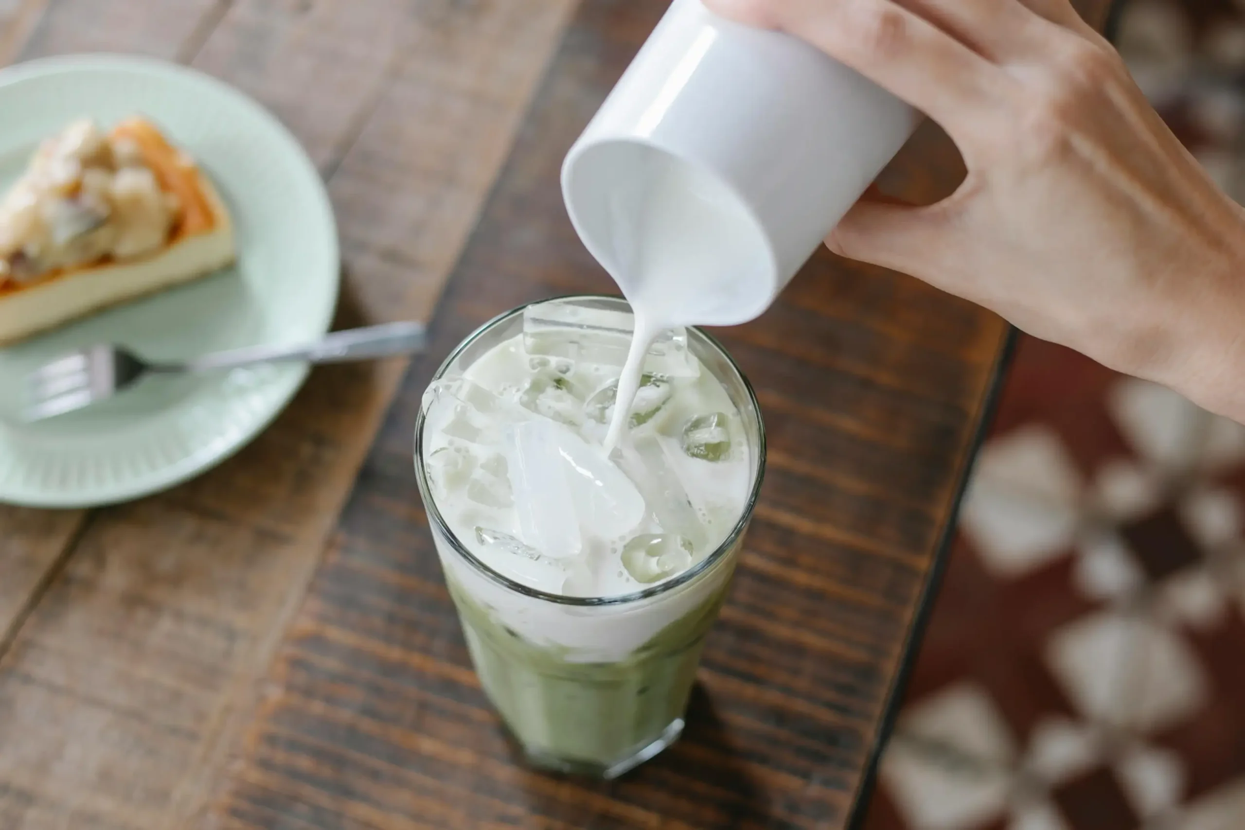 A hand pours fresh milk from a white pitcher into a tall glass of iced matcha tea on a rustic wooden table. In the background, a slice of creamy cheesecake sits on a light green plate, completing the café scene.