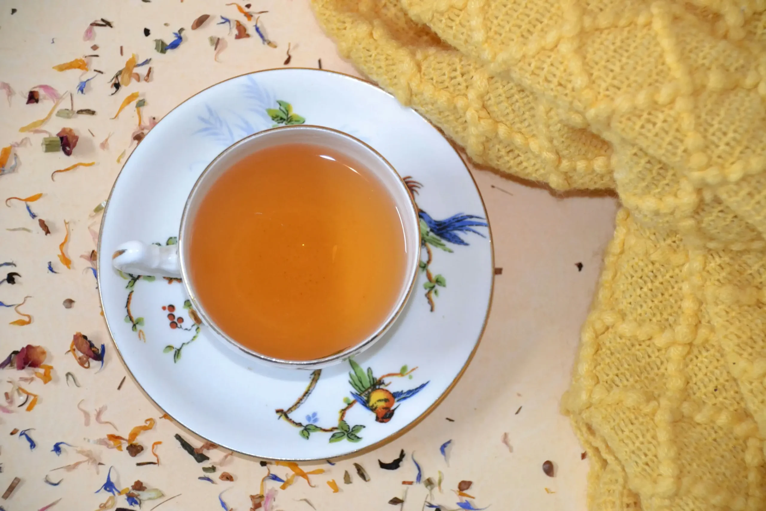 An elegant teacup filled with amber-colored tea rests on a decorative saucer, surrounded by scattered dried flower petals. A cozy, textured yellow knit blanket is draped beside the cup, adding a warm and inviting feel to the scene