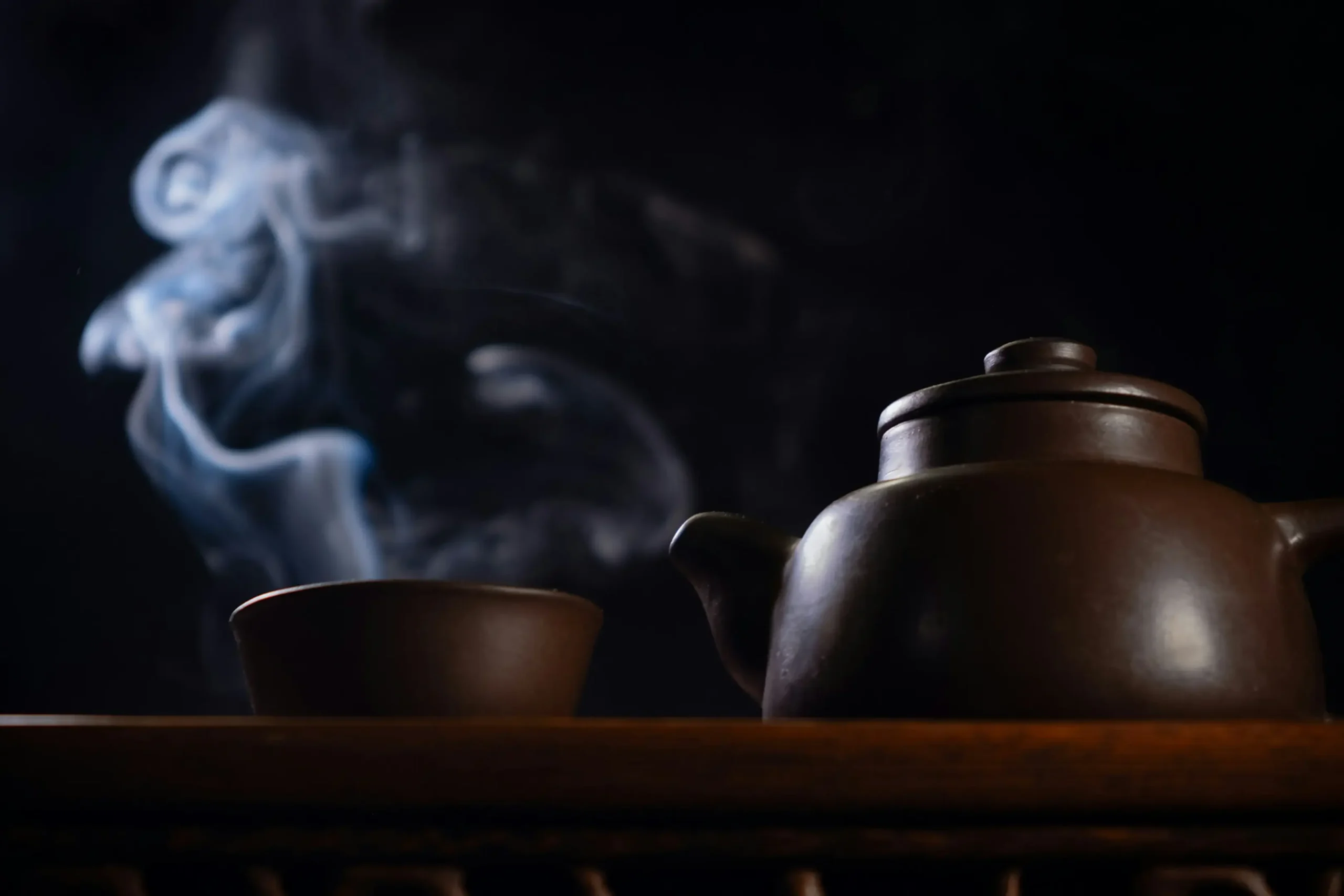 A dark ceramic teapot and a matching cup sit on a wooden surface against a black background. Wisps of white steam curl upward from the cup, creating a serene and moody atmosphere.