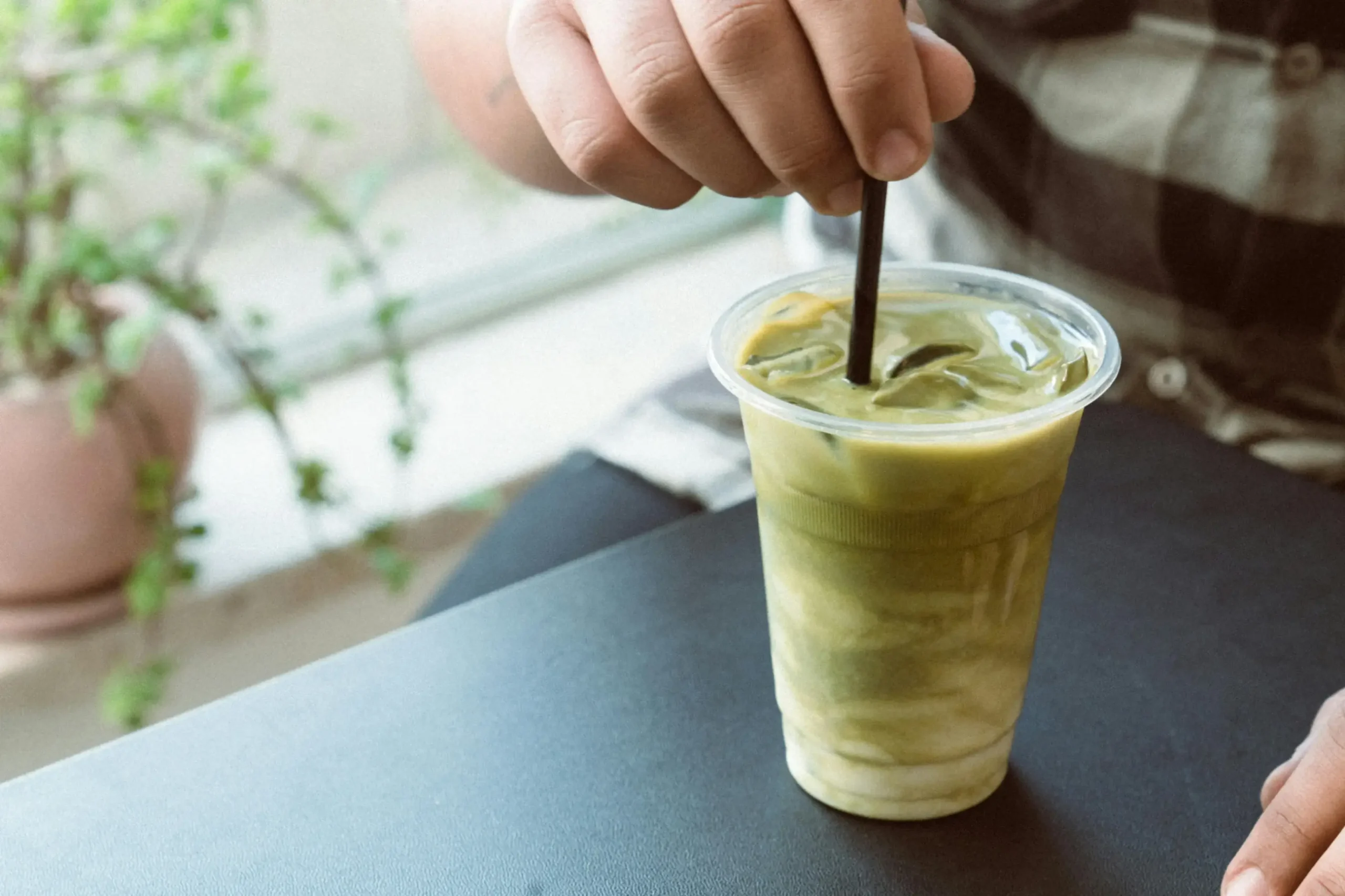 A person uses a black straw to stir an iced matcha latte in a clear plastic cup, blending the vibrant green tea with swirls of milk. The drink sits on a dark table next to a window, with a soft-focus potted plant adding a touch of nature to the background.