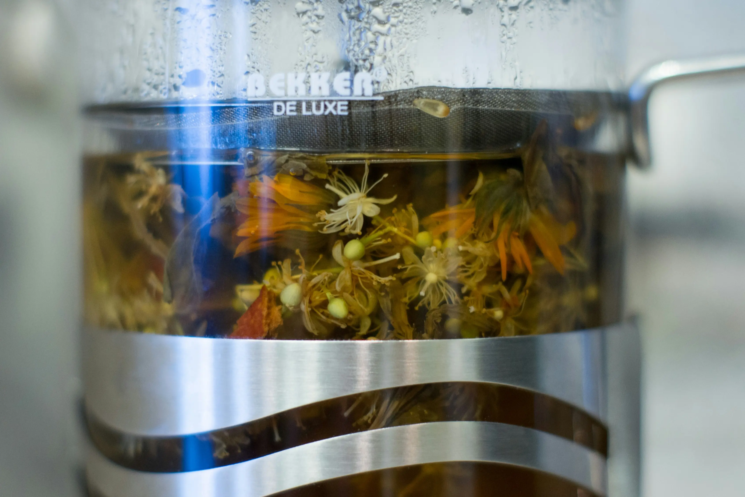 A glass French press is filled with water and a vibrant assortment of steeping botanical ingredients, including yellow and orange flower petals. The mesh filter at the top holds the herbs submerged as the liquid begins to take on a golden hue.