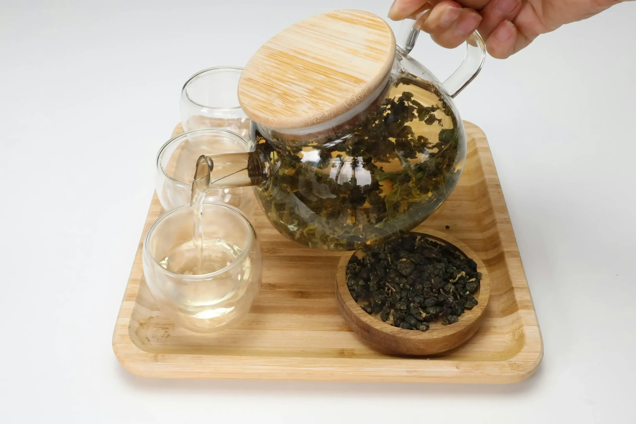 A hand pours pale green tea from a glass teapot with a wooden lid into one of three small glass cups. The set is arranged on a light wooden tray alongside a small bowl of dried, rolled tea leaves.