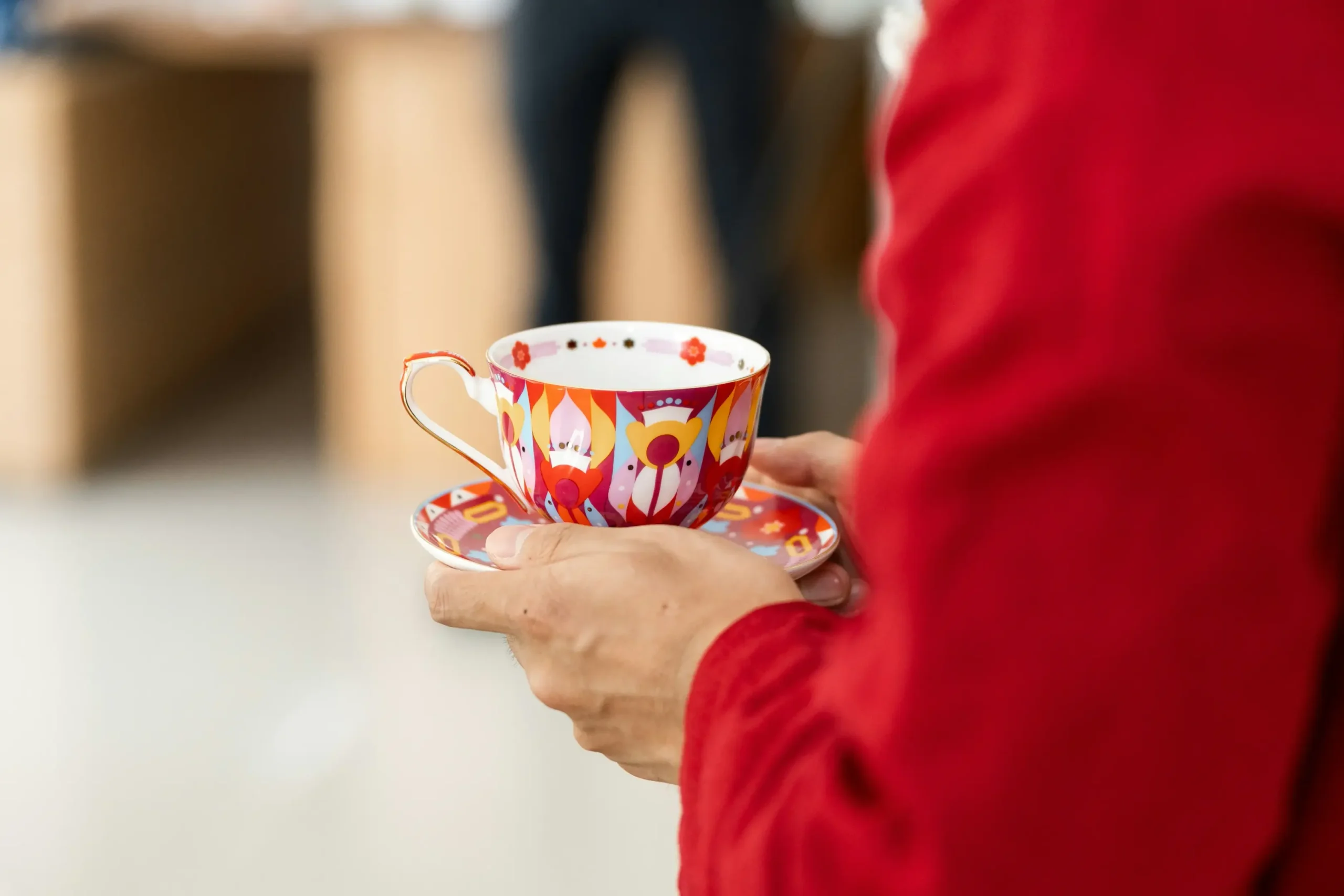 A person wearing a red shirt holds a vibrant, multi-colored teacup and matching saucer with both hands. The ornate porcelain features a bold, symmetric pattern in shades of red, yellow, and blue against a blurred indoor background.