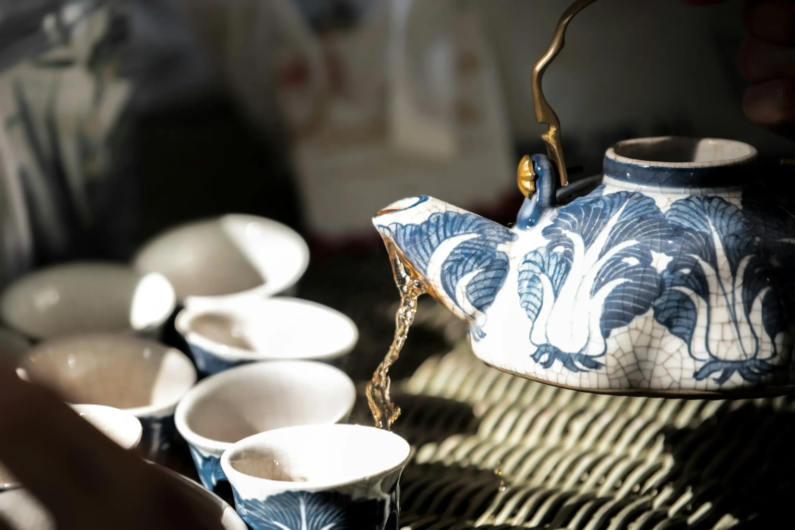 A decorative blue and white ceramic teapot pours amber tea into a matching cup, captured in a moment of fluid motion. The scene features a shallow depth of field, highlighting the intricate floral patterns of the pottery against a soft, sunlit background.