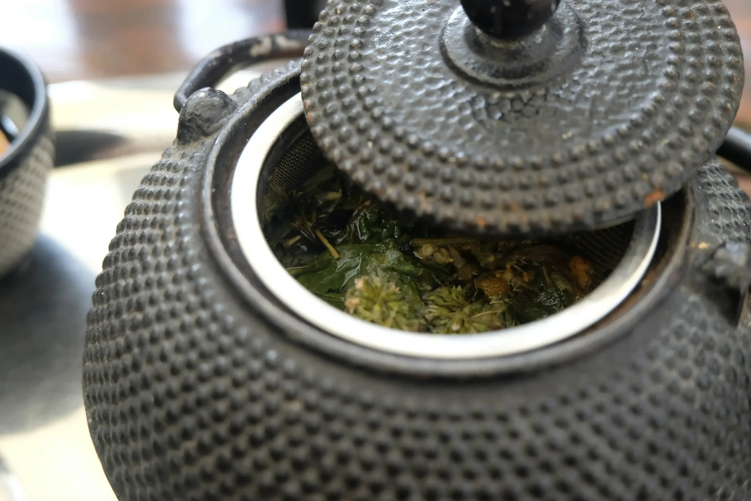 A textured black cast-iron teapot sits with its lid slightly ajar, revealing a metal infuser filled with steeping green tea leaves. The close-up shot highlights the intricate hobnail pattern on the pot's surface and the vibrant, wet herbs inside.