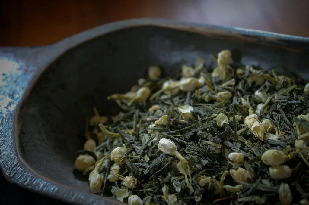 A rustic wooden bowl is filled with a mixture of dark green tea leaves and small, dried jasmine flower buds. The image features a shallow depth of field, highlighting the delicate textures of the botanicals against a dark, moody background.