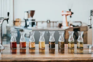 Eight glass carafes filled with various colorful tea infusions and botanical ingredients are lined up neatly on a wooden counter. In the blurred background, professional coffee and tea brewing equipment sits in a bright, modern cafe setting.