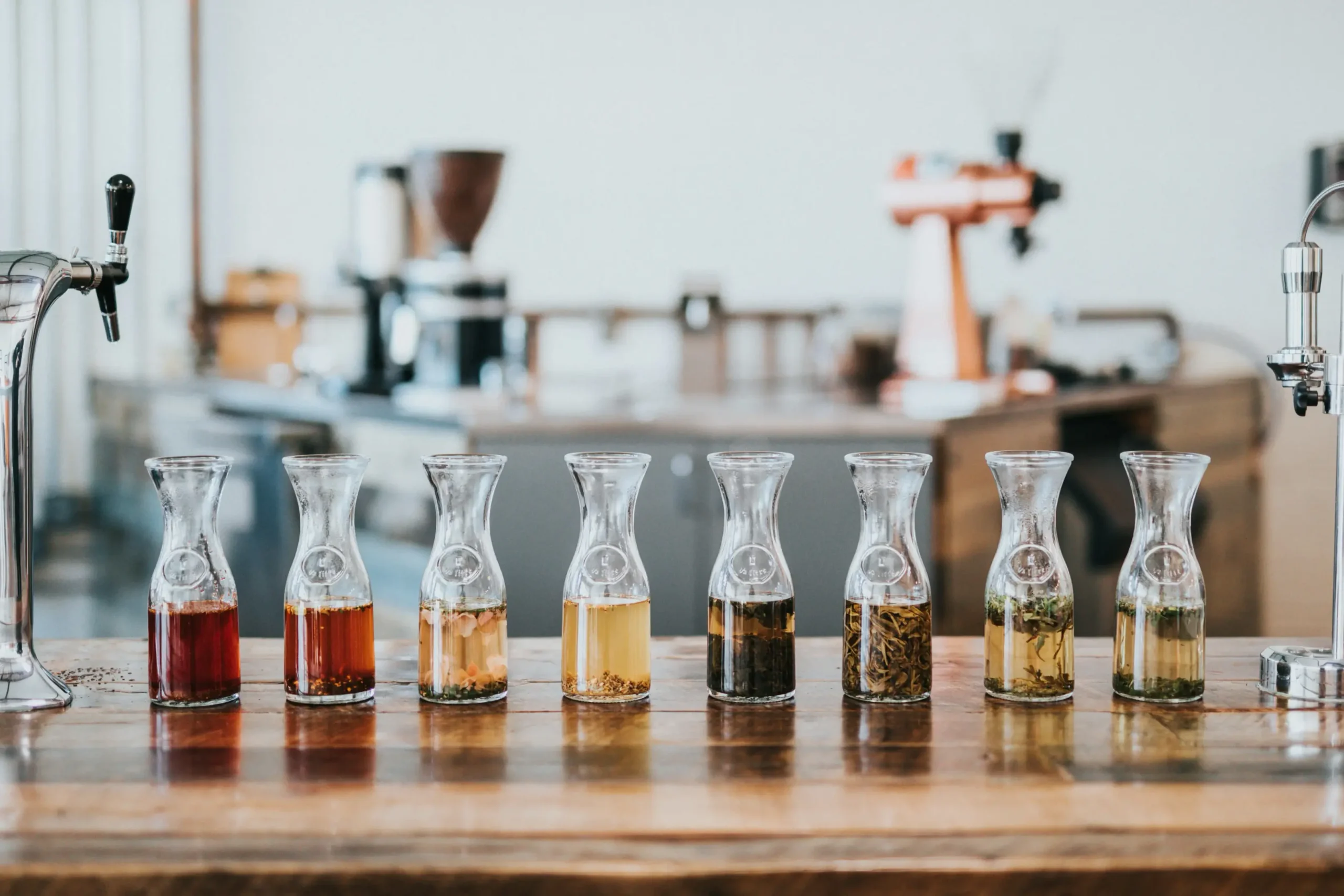 Eight glass carafes filled with various colorful tea infusions and botanical ingredients are lined up neatly on a wooden counter. In the blurred background, professional coffee and tea brewing equipment sits in a bright, modern cafe setting.
