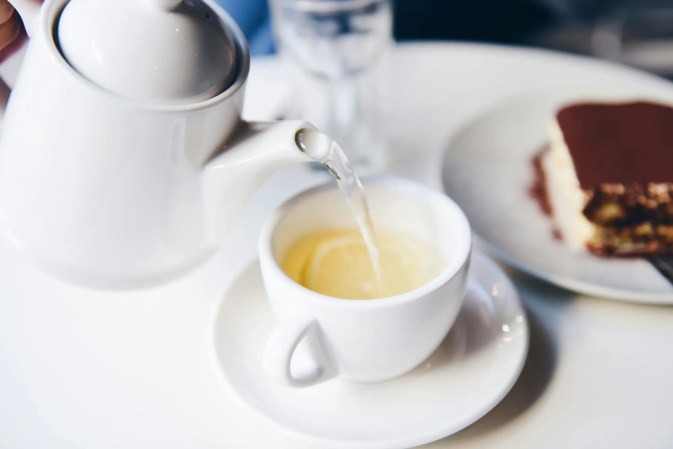 A white teapot pours a clear liquid into a matching teacup containing fresh lemon slices. In the blurred background, a slice of tiramisu rests on a plate alongside an empty glass on a white table.