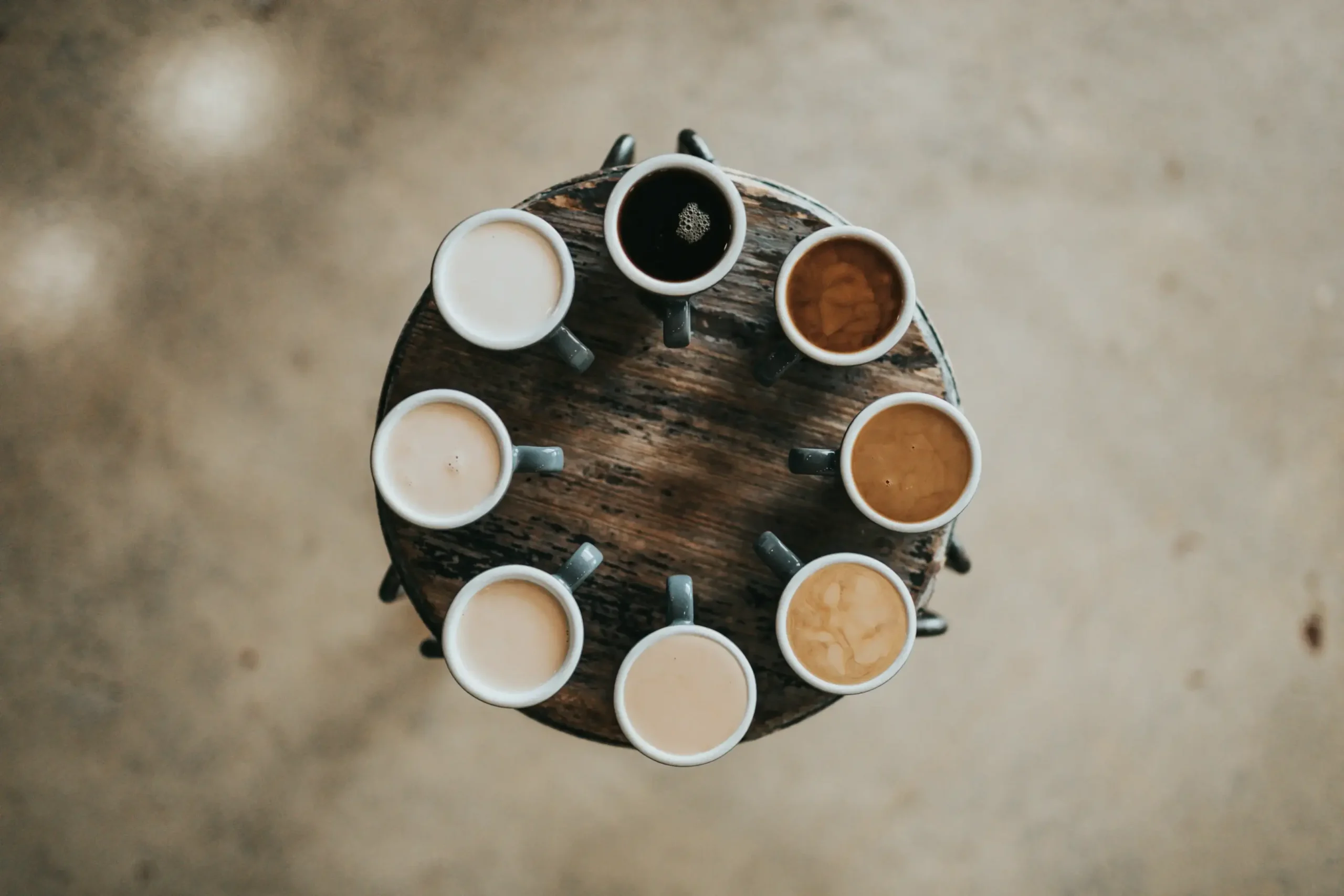 A top-down view shows eight mugs of coffee arranged in a circle on a rustic wooden stool, displaying a gradient of colors from dark black to creamy light tan. The composition is centered against a soft, neutral-toned background, highlighting the variety of coffee blends and milk ratios.