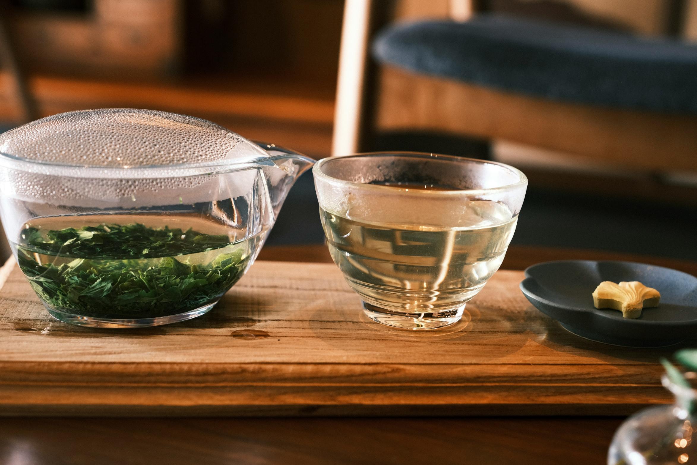 A clear glass teapot sits on a wooden tray, filled with water and vibrant green tea leaves that are steeping inside. Next to it, a small glass cup holds the pale brewed tea, accompanied by a tiny yellow sweet on a dark petal-shaped plate.