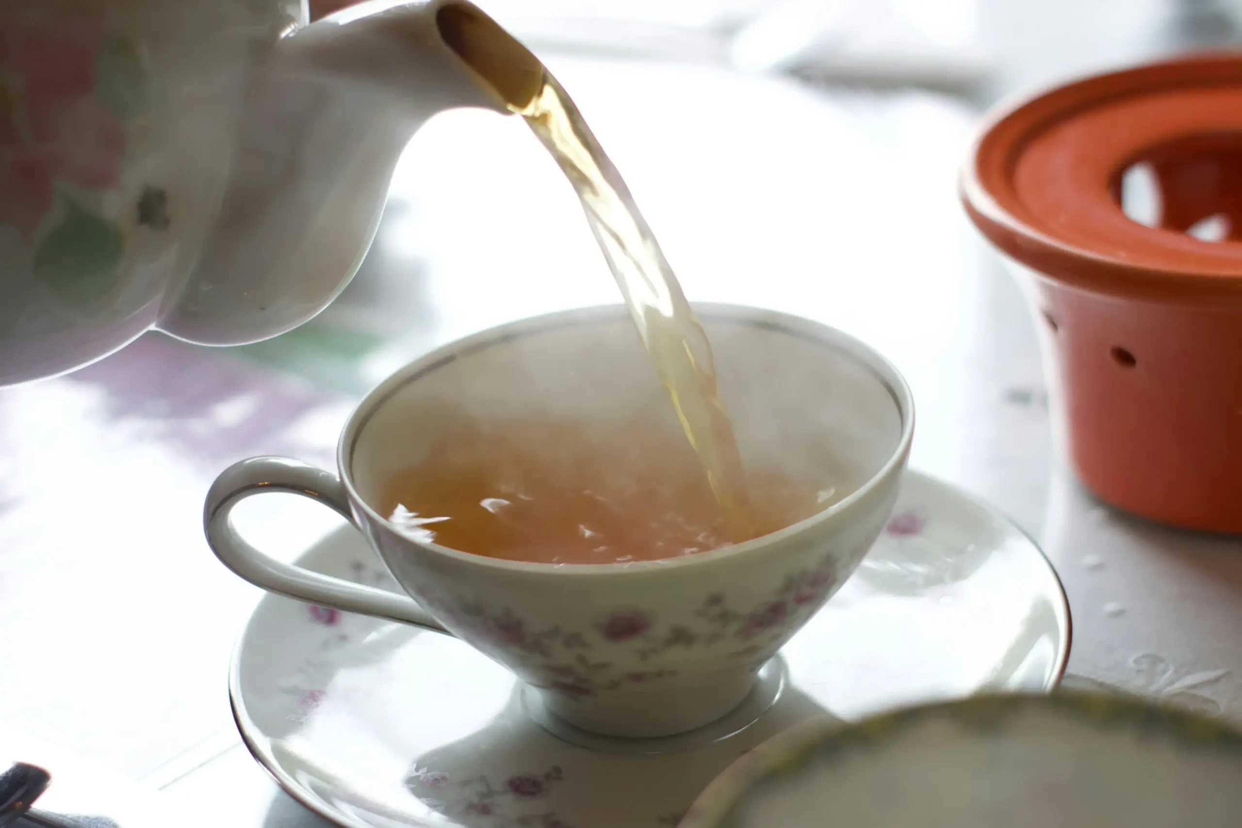 A steady stream of amber tea pours from a floral-patterned teapot into a matching delicate teacup. Light steam rises from the cup, which rests on a saucer against a soft, brightly lit background.