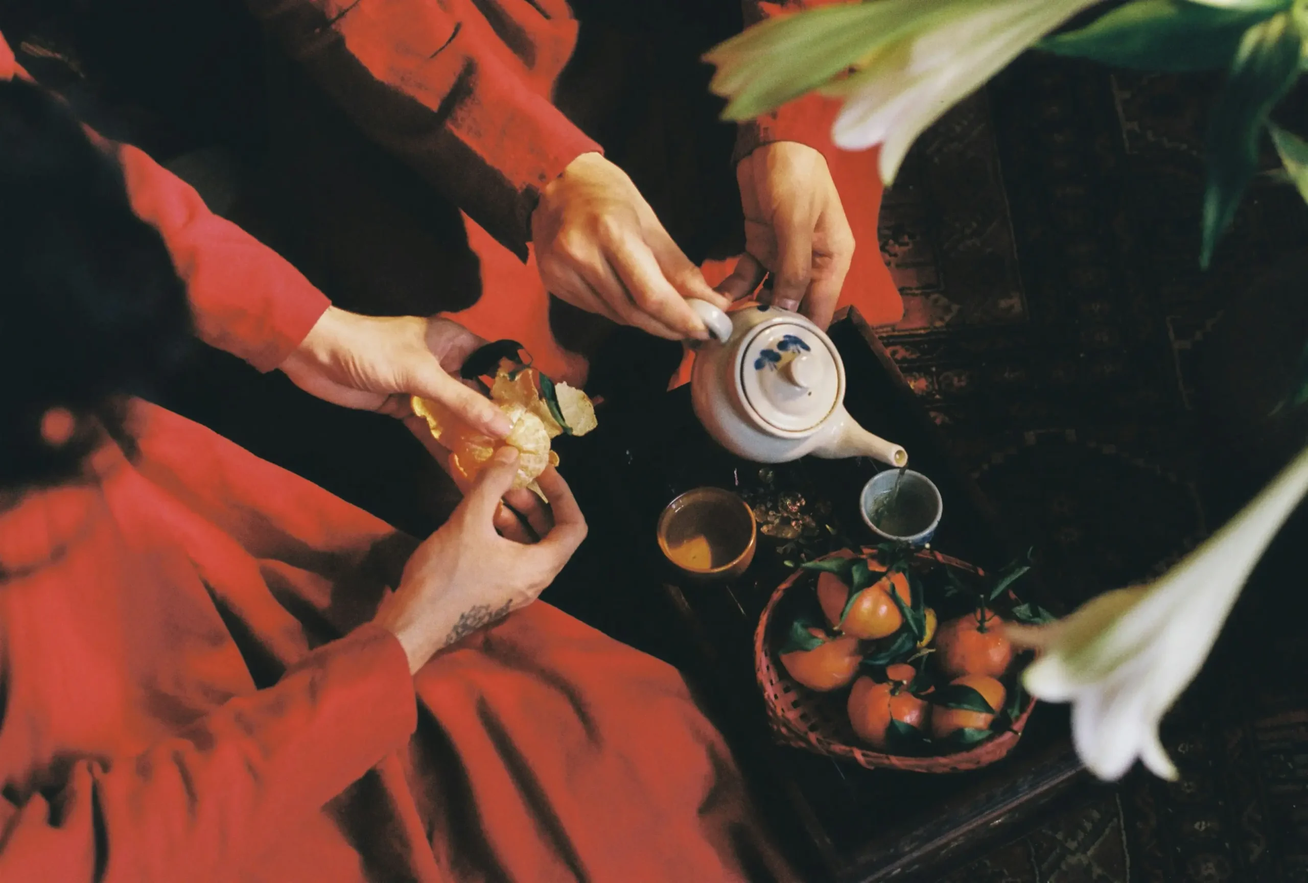 Two people wearing vibrant red garments share a traditional moment of tea and fruit over a patterned rug. One person carefully pours tea from a ceramic pot while the other peels a fresh tangerine.
