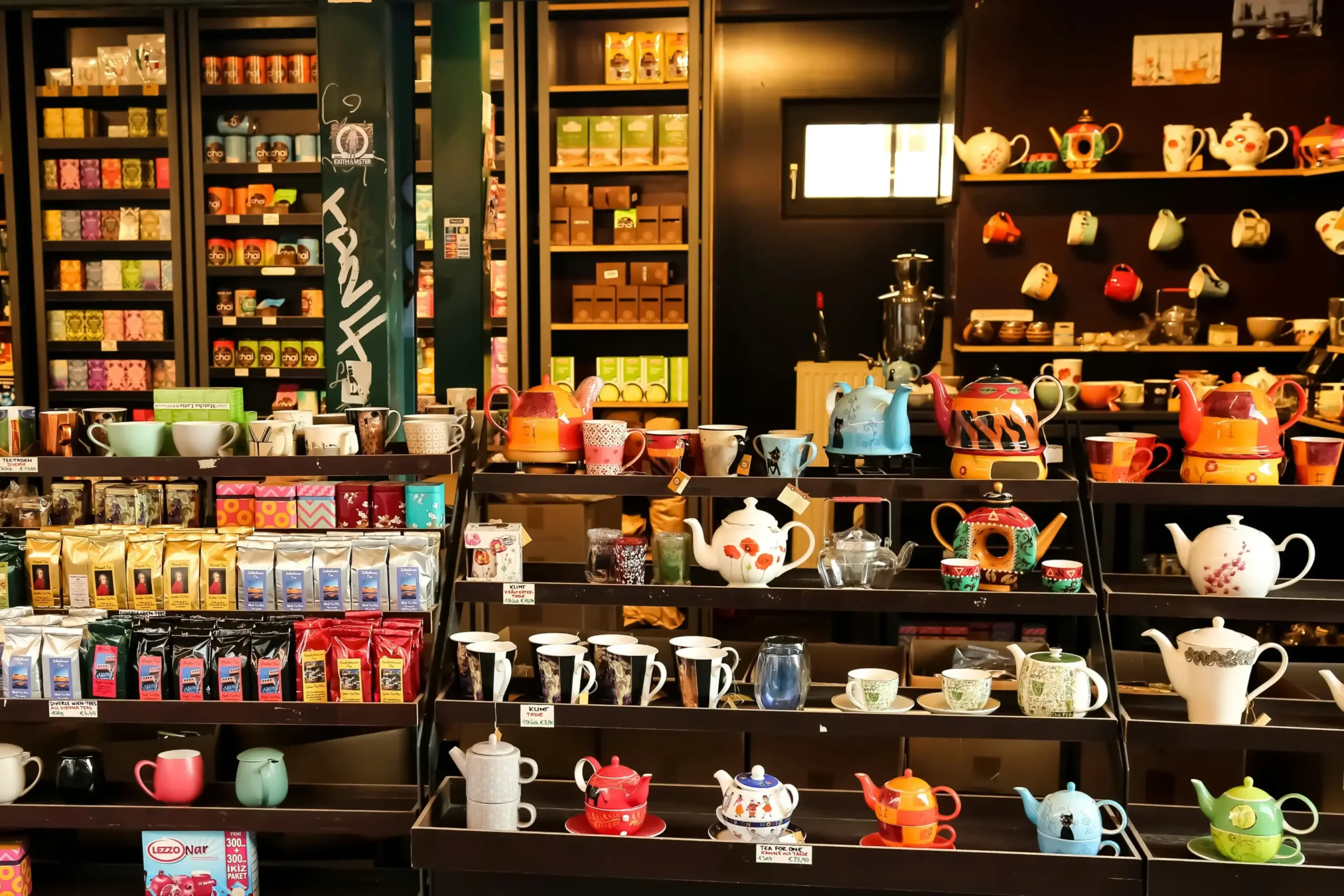 This image showcases a vibrant shop filled with an extensive collection of tea-related products, featuring shelves lined with colorful tea tins and bags. The foreground highlights a diverse display of intricately designed teapots and matching mugs arranged on tiered black stands.