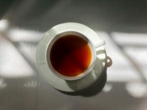 A white ceramic cup filled with dark amber tea sits centered on a matching saucer against a clean white surface. Soft morning light and shadows from a nearby window create a high-contrast, minimalist pattern across the scene.