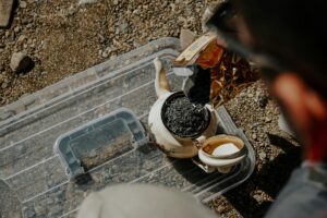A person pours loose tea leaves from a gold-lined bag into a vintage, floral-patterned teapot resting on a clear plastic lid. The scene takes place outdoors on a rugged, gravelly ground under bright sunlight.