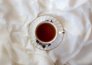 A delicate porcelain teacup filled with dark tea sits centered on a matching floral saucer. The set is nestled within the soft, rippling folds of a crisp white fabric, creating a clean and serene aesthetic.