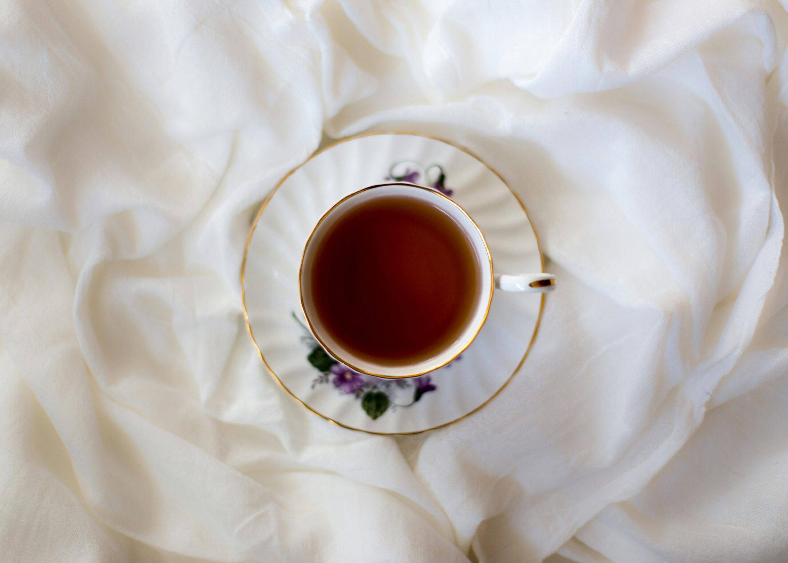 A delicate porcelain teacup filled with dark tea sits centered on a matching floral saucer. The set is nestled within the soft, rippling folds of a crisp white fabric, creating a clean and serene aesthetic.