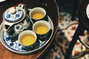A blue and white floral teapot sits on a matching tray alongside three small cups filled with golden tea. The set is positioned on a dark wooden table, with a glimpse of decorative mosaic tiling visible on the floor below.