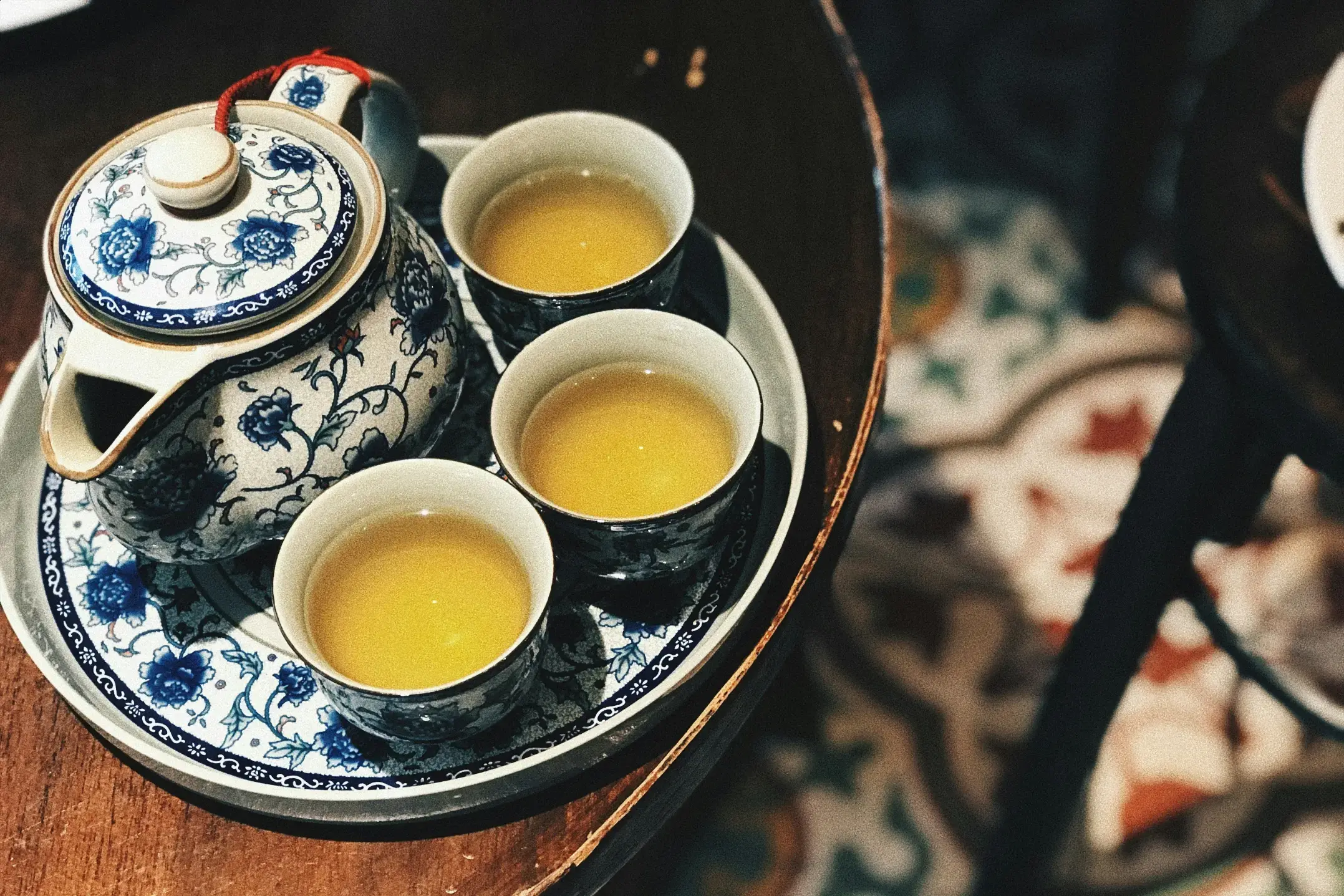 A blue and white floral teapot sits on a matching tray alongside three small cups filled with golden tea. The set is positioned on a dark wooden table, with a glimpse of decorative mosaic tiling visible on the floor below.