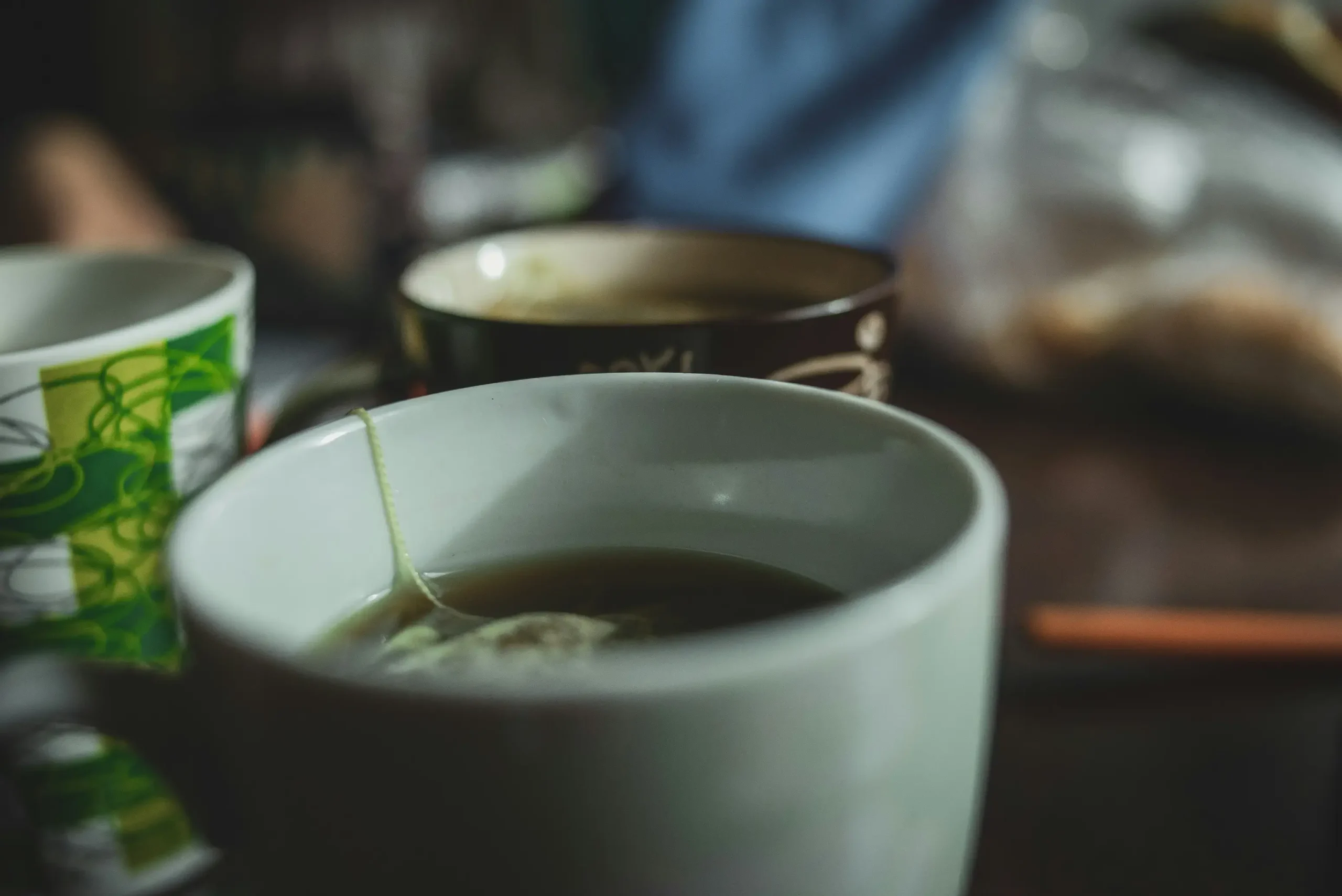 Several mugs filled with dark liquid sit on a table, with a tea bag string hanging over the edge of the closest white cup. The scene is captured with a shallow depth of field, creating a moody and intimate atmosphere.