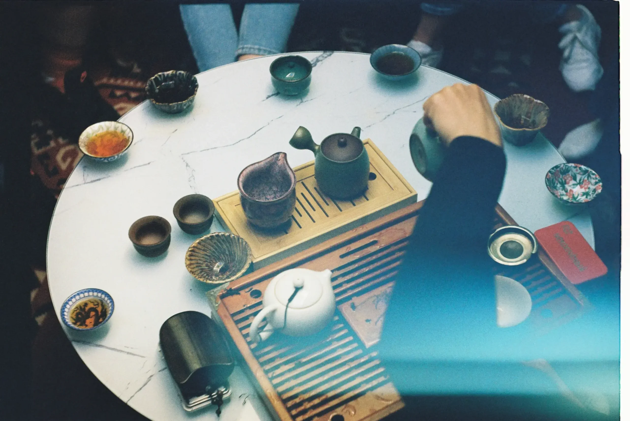 A group of people gathers around a marble-topped table for a traditional tea ceremony, featuring various teapots, small cups, and wooden tea trays. The scene is captured with a nostalgic, film-like aesthetic, including a prominent blue light leak across the foreground.
